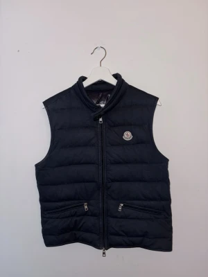 Mörk blå dunväst från Moncler - Snygg mörk blå dunväst från Moncler med quiltad design och hög krage. Västen har två dragkedjeförsedda fickor framtill och Monclers ikoniska logga på bröstet. Perfekt att slänga över en hoodie eller tröja för extra värme. Ps 2500 snabb affär 