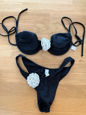 Svart bikini med vita blommor  - Snygg svart bikini från shein med vita blommor som dekoration på både överdel och underdel. Överdelen har knytband och vadderade kupor, medan underdelen har en klassisk passform. Perfekt för stranden eller poolen!