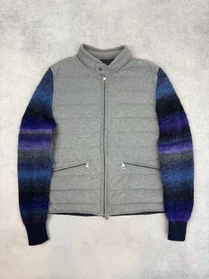 Grå quiltad jacka från Moncler - Unik grå quiltad jacka från Moncler med stickade ärmar i blå, svart och lila toner. Jackan har dragkedja framtill, ståkrage och två snedställda fickor med dragkedja. Perfekt för dig som vill sticka ut med coola färgövergångar på ärmarna.