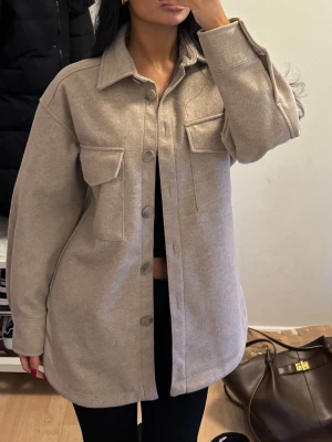 Beige overshirt med fickor - Stilren beige overshirt i mjukt material med klassisk krage och stora bröstfickor med lock. Rak passform och knäppning framtill. Perfekt lager-på-lager-plagg för en avslappnad look.