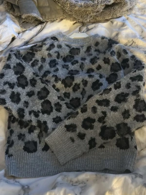 Grå leopardmönstrad stickad tröja - Säljer en grå stickad tröja med svart leopardmönster. Tröjan har rund halsringning, långa ärmar och ribbade muddar vid ärmslut och nederkant. Perfekt för dig som vill ha en trendig och mysig look.