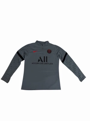 Grå PSG träningströja från Nike - Snygg grå långärmad träningströja från Nike med Paris Saint-Germain-logga och röd Nike swoosh på bröstet. Tröjan har halv dragkedja och svart tryck med 'Accor Live Limitless' på framsidan. Perfekt för fotbollsträning eller sport.