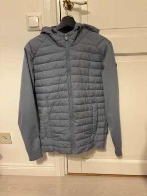 Ljusblå pufferjacka med huva - Snygg ljusblå pufferjacka med quiltad front och huva. Jackan har dragkedja framtill, ribbade muddar vid ärmslut och nederkant samt släta ärmar. Perfekt för kalla dagar och ger en clean look.