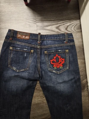Dsq2 jeans med röd lönnlöv HELG REA - Mörkblå jeans från Dsq2 med snyggt sliten tvätt och kontrastsömmar. Bakfickan har en stor röd broderad lönnlöv och siffrorna 1995. Klassisk femficksmodell med läderpatch i midjan. Perfekta för dig som gillar streetwear och detaljer. Har mått som jag kan skicka 