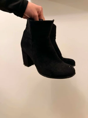 Svarta mockastövletter med klack - Säljer ett par svarta stövletter i mocka-look med hög, bred klack och rundad tå. Skorna har en stilren och enkel design som passar till många outfits. Perfekta för dig som gillar klassiska boots med lite höjd.