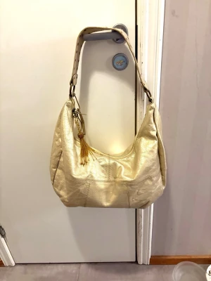 Guldig axelremsväska i skinn - Ny snygg  guldig handväska i mjukt skinnl med bred axelrem och dekorativa nitar o tofsdetalj.  Rymlig modell 40*28 cm.  