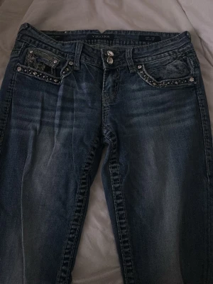 blå lowwaist bootcut jeans från vigoss - Säljer ett par blå bootcut lowwaist jeans från Vigoss med snygga slitningar och dekorativa nitar på fickorna. Jeansen har en klassisk femficksmodell och coola broderade detaljer på bakfickorna. Perfekta för dig som vill ha ett par jeans med lite extra edge. storlek 5/6 och längd 33. Pris kan diskuteras