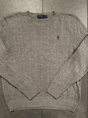 Grå kabelstickad tröja Polo Ralph Lauren - Kabelstickad grå tröja från Polo Ralph Lauren med rund halsringning och klassisk broderad logga på bröstet. Tröjan har långa ärmar och ribbade muddar vid ärmslut och nederkant. Perfekt för lager-på-lager och stilren look.