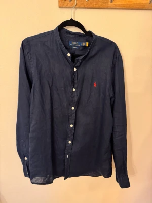 Mörkblå linneskjorta från Polo Ralph Lauren - Klassisk mörkblå skjorta i linne från Polo Ralph Lauren med vit knappslå och den ikoniska röda logobrodyr på bröstet. Skjortan har lång ärm, normal passform och en stilren, tidlös look som passar till det mesta.