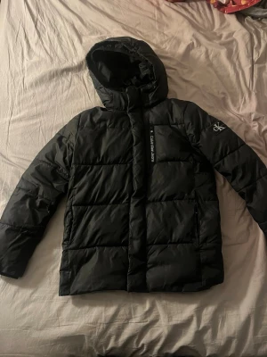Svart pufferjacka från Calvin Klein - Snygg svart pufferjacka från Calvin Klein Jeans med huva och dragkedja framtill. Jackan har logga på ärmen och insidan samt stora quiltade partier. Perfekt för kalla dagar och ger en modern streetwear-look.