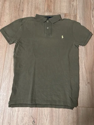 Olivgrön piké från Polo Ralph Lauren - Olivgrön pikétröja från Polo Ralph Lauren med klassisk krage, två knappar och gul broderad logga på bröstet. Tillverkad i mjuk bomull och har korta ärmar för en clean och tidlös stil.