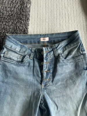 Ljusblå jeans från Nelly med knappar - Snygga ljusblå jeans från Nelly med klassisk femficksmodell och knappgylf. Jeansen har en rak passform och är tillverkade i mjukt denimtyg. Perfekta för en avslappnad och trendig look.