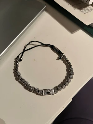 Svart och silverfärgat pärlarmband - Stilrent armband med svarta snören och silverfärgade pärlor i olika former. Armbandet har en rektangulär detalj i mitten med mönster och justerbar knytning. Perfekt accessoar för att lyfta din outfit med en cool touch.