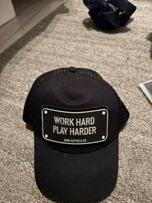 Svart keps John Hatter & Co - Snygg svart keps från John Hatter & Co med texten 'WORK HARD PLAY HARDER' på en metallskylt framtill. Kepsen har böjd skärm och mesh på sidorna och bak för extra ventilation. Perfekt accessoar för en cool och avslappnad stil.