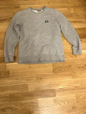 Grå sweatshirt från J.Lindeberg - En stilren grå sweatshirt från J.Lindeberg med rund halsringning och diskret logga broderad på bröstet. Tröjan har långa ärmar och ribbade muddar vid ärmslut och nederkant. Perfekt för en avslappnad och snygg look. Har små defekter vid ena armen( bild 3) men syns inte så mycket vad användning.