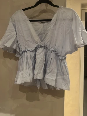 Ljusblå volangblus från Zara Basic - Supersöt ljusblå blus från Zara Basic med v-ringning, volangdetaljer och lös passform. Blusen har korta, vida ärmar och rynkade sömmar i midjan som ger en flowy look. Perfekt att styla med jeans eller kjol för en trendig vibe. Säljs inte längre deflr högre pris liten defekt som syns på bild men försvinner i tvätt