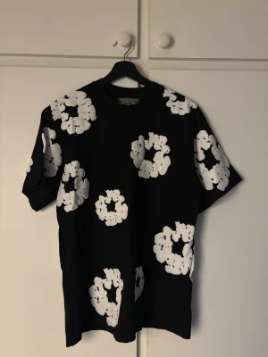 Svart blommig t-shirt från Denim Tears - Svart t-shirt från Denim Tears med stora vita blommönster över hela tröjan. Klassisk passform med rund hals och korta ärmar. Perfekt för dig som vill sticka ut med en unik och trendig look. Storlek S men passar M 