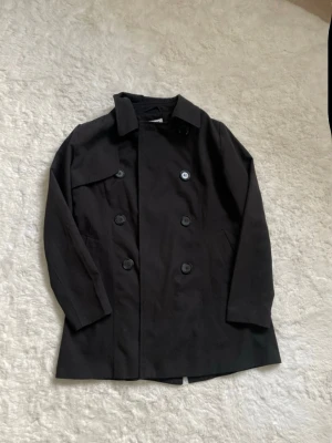 Svart klassisk trenchcoat - Stilren svart trenchcoat med dubbelknäppning och klassisk krage. Knappt använt. Kommer dock utan bälte.