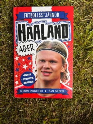 Fotbollsstjärnor: Haaland äger - En färgstark bok om fotbollsstjärnan Erling Haaland, fylld med fakta, statistik och bragder. Omslaget är rött med blå och vita detaljer, och boken är skriven av Simon Mugford och Dan Green. Perfekt för dig som älskar fotboll och vill veta mer om Haalands resa till toppen.