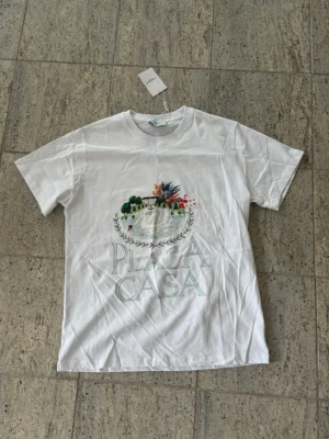 Vit t-shirt med Plaza Casa-tryck - Vit t-shirt med rund hals och tryck framtill som föreställer en villa med palmer och blommor samt texten 'PLAZA CASA' i stora bokstäver. T-shirten har en klassisk passform och är tillverkad i mjukt bomullsmaterial. Perfekt för en avslappnad och trendig look.