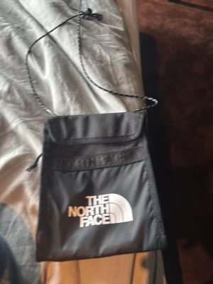 Svart axelväska från The North Face - Snygg svart axelväska från The North Face med vit logga framtill. Väskan har en justerbar snodd för att bäras över axeln och ett lock med kardborrestängning. Tillverkad i slitstarkt syntetmaterial och har en minimalistisk, fyrkantig design.