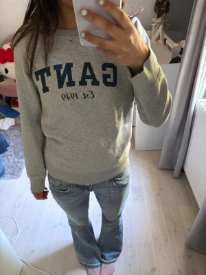Grå sweatshirt från GANT - Säljer en grå sweatshirt från GANT med blå logga och texten 'Est. 1949' på bröstet. Tröjan har rund halsringning, långa ärmar och mjukt, borstat foder på insidan. Perfekt för en avslappnad och stilren look.