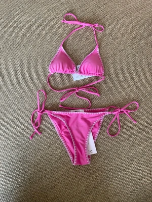 Rosa bikini med knyt från Gina Tricot - Supersöt rosa bikini från Gina Tricot med ljusrosa detaljer längs kanterna. Klassisk triangelmodell med knyt både i ryggen, nacken och på sidorna av bikinitrosan. Perfekt för strandhäng och poolparty! Helt ny aldrig använd. Hör av er om ni vill ha mått eller fler bilder! 
