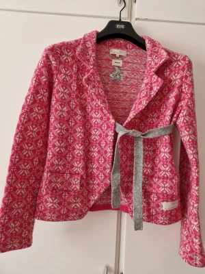 Rosa mönstrad cardigan från Odd Molly - Superfin rosa cardiganjacka från Odd Molly med vitt mönster, grått knytband i midjan och broderade detaljer på ryggen. Jackan har krage, två fickor fram och är stickad i mjukt material. Perfekt för dig som gillar färg och unika detaljer.