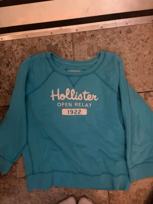 Turkos sweatshirt från Hollister - Turkos sweatshirt från Hollister med tryck 'Hollister Open Relay 1922' på bröstet. Tröjan har rund halsringning, långa ärmar och är gjord i mjukt bomullsmaterial
