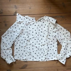 Vit prickig blus från KappAhl - Supersöt vit blus från KappAhl med svarta prickar och volangdetaljer framtill. Blusen har långa ärmar med knappar vid ärmsluten och en rund halsringning. Perfekt för dig som gillar en lekfull och trendig stil.