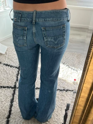 Diesel jeans low waist med vida ben - Säljer ett par blå low waist jeans med vida ben och dubbla knappar i midjan. Jeansen har klassiska fickor fram och bak samt bälteshällor. Tillverkade i denim med en snygg tvättad look, perfekta för en trendig och avslappnad stil. Defekt på sista bilden, syns inte på(går att sy)