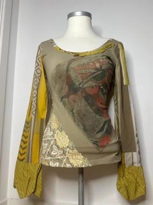 Gul mönstrad långärmad topp - Unik långärmad topp i beige och gula toner med blandade mönster och patchwork-detaljer. Toppen har vida ärmar, broderade partier och en cool mix av grafiska och blommiga inslag. Perfekt för dig som gillar statement-plagg och kreativ stil. Mått: Armhåla till armhåla: 41 cm, Längd: 42 cm och Ärmlängd: 61 cm
