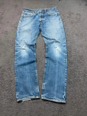 Levi's 501 jeans vintage - Klassiska blå jeans från Levi's med rak passform och slitningar på knäna. Jeansen har fem fickor, knappgylf och tydliga slitdetaljer för en cool vintagekänsla. Perfekta för dig som gillar en avslappnad och tidlös stil. De är 32/32 i storlek hör gärna av dig om du har några frågor☺️