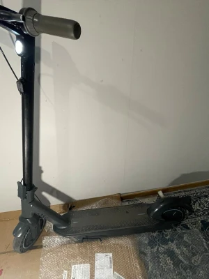 Svart hopfällbar elsparkcykel - Elektrisk sparkcykel i svart med grå handtag och robust ram i metall. Utrustad med stora hjul med blå ringdetaljer, bred fotplatta med halkskydd och framlykta. Styret är justerbart och modellen är hopfällbar för enkel transport och förvaring.