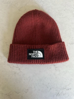 Rostbrun mössa från The North Face - Säljer en snygg rostbrun ribbstickad mössa från The North Face. Mössan har uppvikt kant med en svartvit logotyp framtill. Tillverkad i 97% akryl, 1% elastan och 2% andra fibrer. Unisex och one size, perfekt för kalla dagar.
