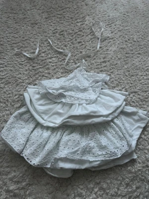 Vit volang topp med spetsdetaljer - Superfin vit bikini med volanger och spetsdetaljer. Toppen har knytband i nacken och dekorativa broderade hålmönster. Perfekt för dig som vill ha en söt och unik look på stranden.
