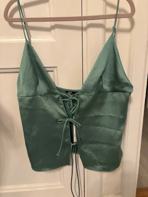 Grön satintopp med snörning från Zara - Supersnygg grön satintopp från Zara med tunna axelband och djup v-ringning. Toppen har snörning framtill som ger en trendig look och är perfekt för att styla med jeans eller kjol. Materialet är glansigt och känns lyxigt mot huden.