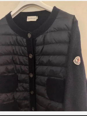 Svart cardiganjacka från Moncler - Storlek L men sitter om S/M !!!!! Stilren svart cardiganjacka från Moncler med quiltad framsida, stickade ärmar och fyra framfickor. Jackan har rund halsringning, knappstängning och Moncler-logga på ärmen. Materialmix av dun och ull ger både värme och snygg look.