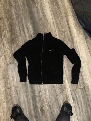 Svart kabelstickad kofta Polo Ralph Lauren - Kabelstickad svart kofta från Polo Ralph Lauren med dragkedja framtill och hög krage. Klassisk vit logga broderad på bröstet och två fickor fram. Ribbstickade muddar vid ärmslut och nederkant. Perfekt för en stilren och avslappnad look.