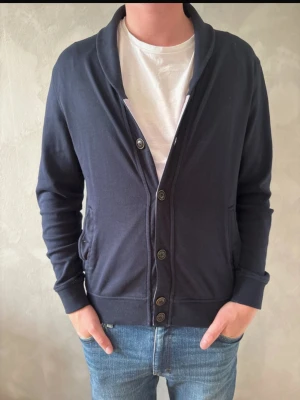 Mörkblå cardigan med knappar - Mörkblå cardigan med avslappnad passform och knäppning framtill. Tröjan har två fickor på sidorna och mjukt material, perfekt att slänga över en t-shirt. Snygg krage som ger en stilren look.