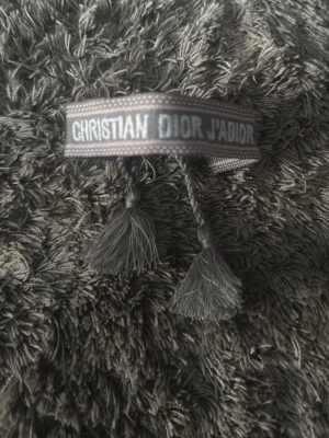 Lätt rosa, Grått tygarmband Christian Dior - Snyggt grått tygarmband från Christian Dior med texten 'CHRISTIAN DIOR J'ADIOR' broderad i vitt. Armbandet har flätade band och tofsar i ändarna för en cool och trendig look. Perfekt accessoar för att lyfta din outfit.