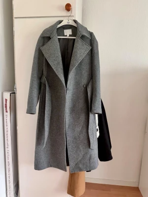 Grå ullrock från H&M, stl XS - Stilren grå ullrock från H&M i storlek XS. Rocken har breda slag, dolda fickor och stängs med dragkedja. Perfekt för kyliga dagar och ger en snygg siluett. Fodrad insida för extra komfort.