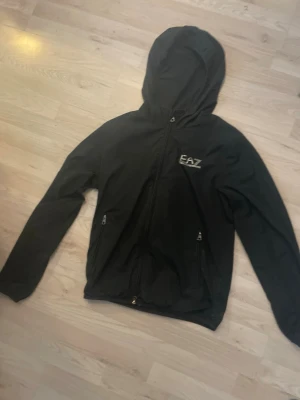 Svart vindjacka från EA7 Emporio Armani - Snygg svart vindjacka från EA7 Emporio Armani med huva och dragkedja framtill. Nypris 1700kr. Jackan har två fickor med dragkedja och även en innerficka. Perfekt för blåsiga/regniga dagar och även svala sommarkvällar. Den är i väldigt bra skick förutom ett pyttelitet hål på framsidan som du kan se på bild 4. Tveka inte på att höra av dig vid några funderingar!🤩