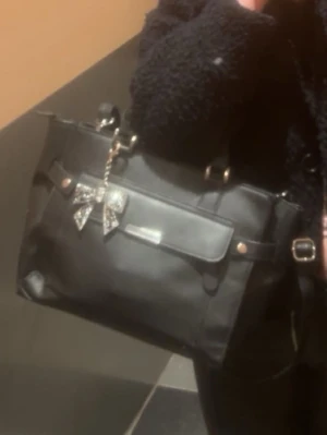 Svart handväska med rosettdetalj - Snygg svart handväska. Väskan har ett ytterfack med ”lock” och två handtag. Söt glittrig rosett.Den är köpt för 400kr Den har en drag kjedja upp till där du kunde stänga väskan. Tyvärr har den gått sönder men går att laga. Därav det billiga priset. Det är ej något som syns när du bär på den osv.