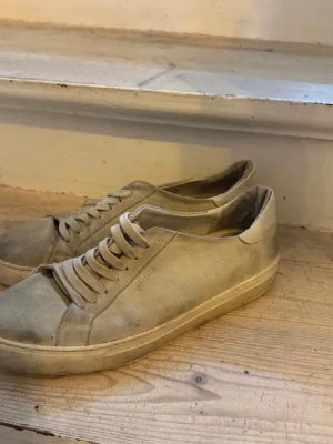 Beige sneakers från Axel Arigato - Stilrena beige sneakers från Axel Arigato med lågt skaft och klassisk siluett. Skorna har ovandel i mocka och matchande snörning. Perfekta för dig som gillar minimalistisk och clean stil.