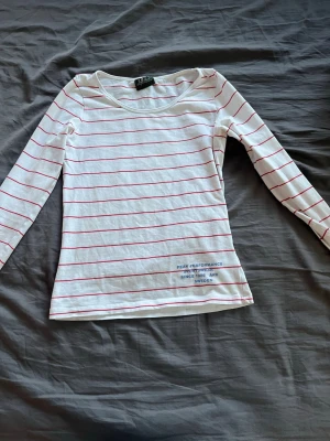 Randig långärmad topp Peak Performance - Vit långärmad topp från Peak Performance med tunna röda ränder och blå broderad text nertill. Toppen har rund halsringning och är tillverkad i mjuk bomull, perfekt för en avslappnad stil.