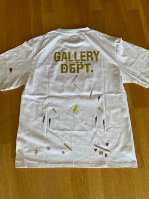 Vit Gallery Dept. t-shirt med tryck - Vit t-shirt från Gallery Dept. med stort tryck på ryggen och färgstänk i olika nyanser. Klassisk passform och rund halsringning. Tillverkad i mjuk bomull, perfekt för en avslappnad och trendig stil.