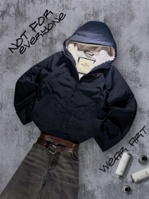 Hollister Jacka med teddyfoder - Svart vindjacka från Hollister med mysigt vitt teddyfoder i huvan och hela insidan. Jackan har dragkedja framtill, två sidofickor och en rymlig huva. Perfekt för kalla dagar när du vill ha både stil och komfort.