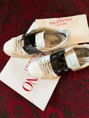 Valentino Garavani vita sneakers - Valentino Garavani med en bred svart lackdetalj över skon. Skorna har klassisk låg modell, vita snören och är tillverkade i läder med en cool kontrast mellan matt och glansigt material.