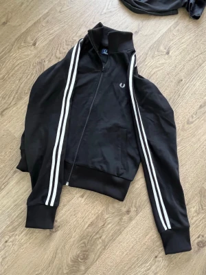 Svart zip-hoodie från Fred Perry - Svart hoodie från Fred Perry med dragkedja och vita dubbla ränder längs ärmarna. Klassisk logga broderad på bröstet. Mjuk och skön bomullsblandning, ribbade muddar och hög krage. Perfekt för en sportig och stilren look. Står storlek 40 men känns mer som 36/38. 